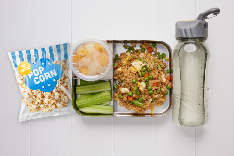 A rectangular metal lunch box containing fried rice, celery sticks, a tub of fruit, a packet of popcorn and a bottle of water