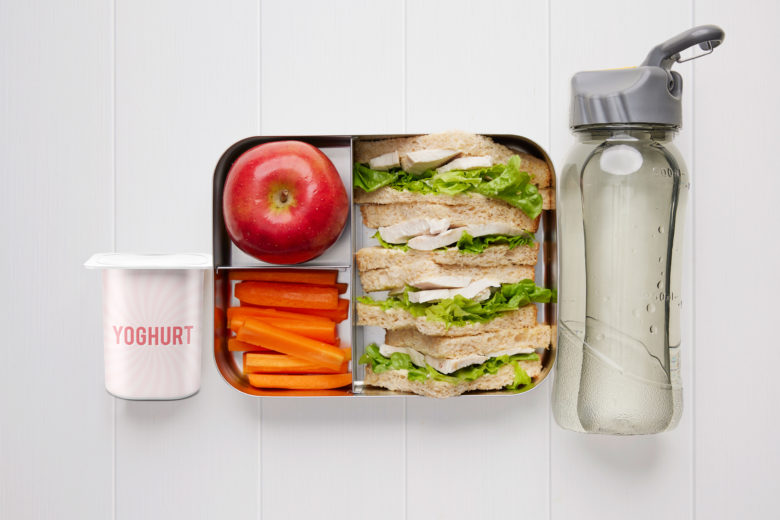 A rectangular metal lunch box containing a chicken and lettuce sandwich, carrot sticks, a red apple, a tub of yoghurt and a bottle of water