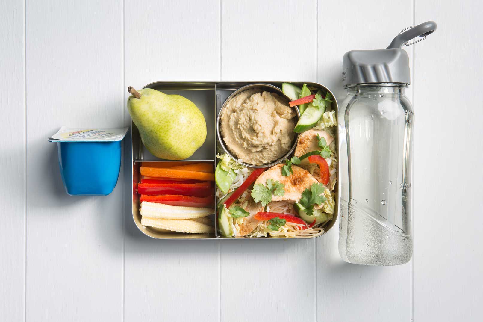 Image of a packed lunch box with soy and ginger chicken noodle salad, vegie sticks with hummus, green pear and a custard tub and bottle of water on the side