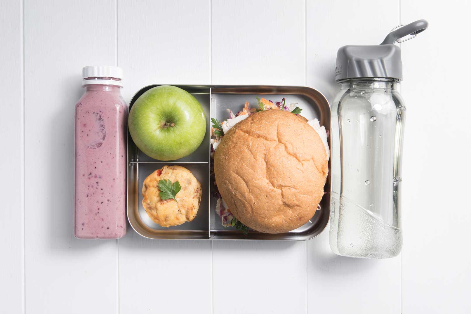 Image of a packed lunch box with chicken and coleslaw roll, easy savoury muffin, green apple and berrylicious smoothie and bottle of water on the side