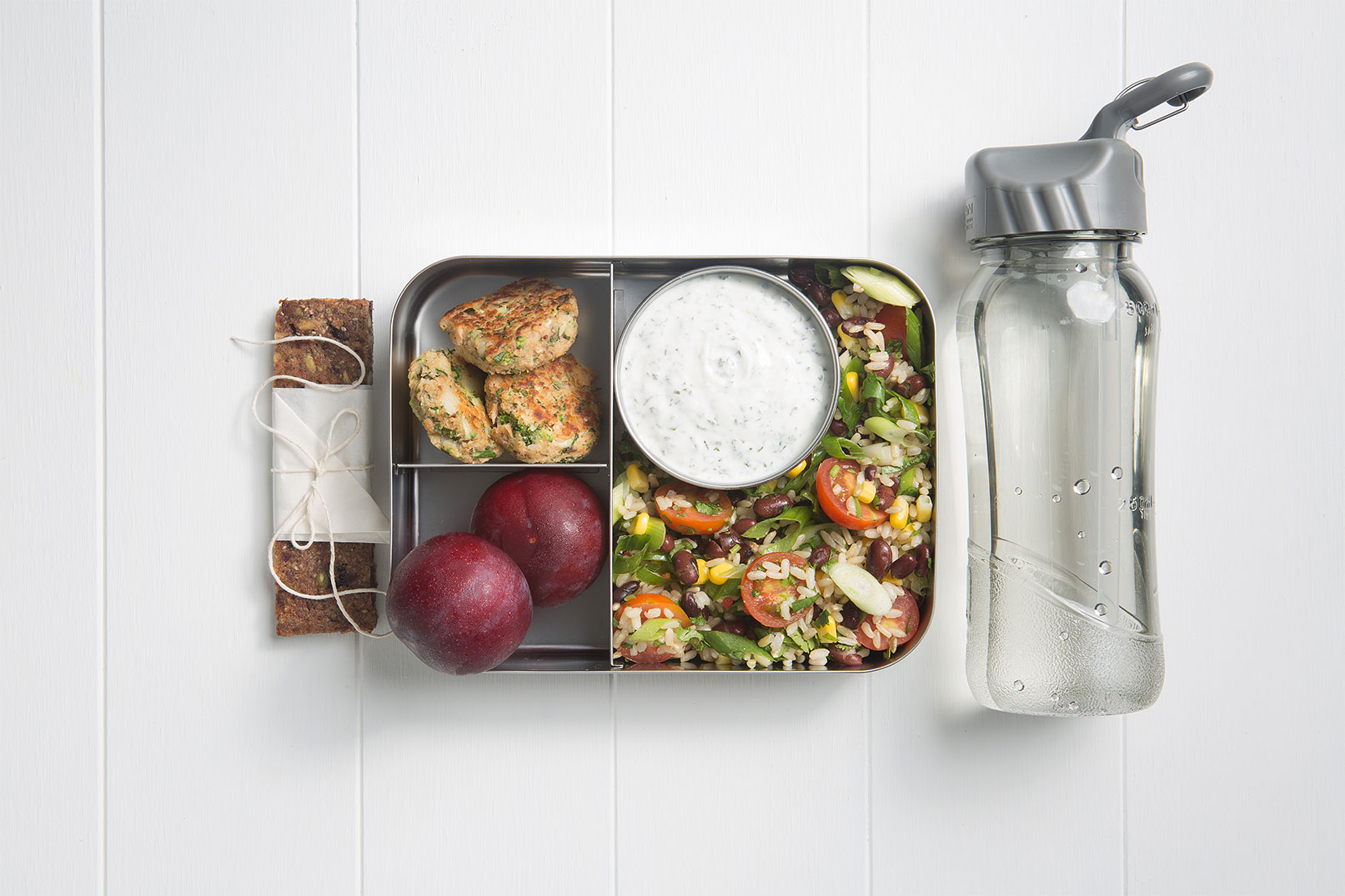 Image of a packed lunch box with zucchini and turmeric salmon patties, rice salad, minty yoghurt dip, two plums, chewy fruit and seed bar and a bottle of water on the side