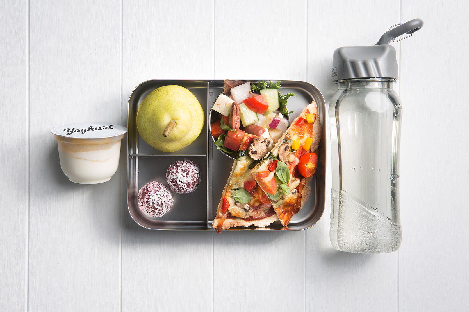Image of a packed lunch box with greek salad, two easy pizza slices, green pear, beetroot and spinach bliss balls, yoghurt tub and a bottle of water on the side
