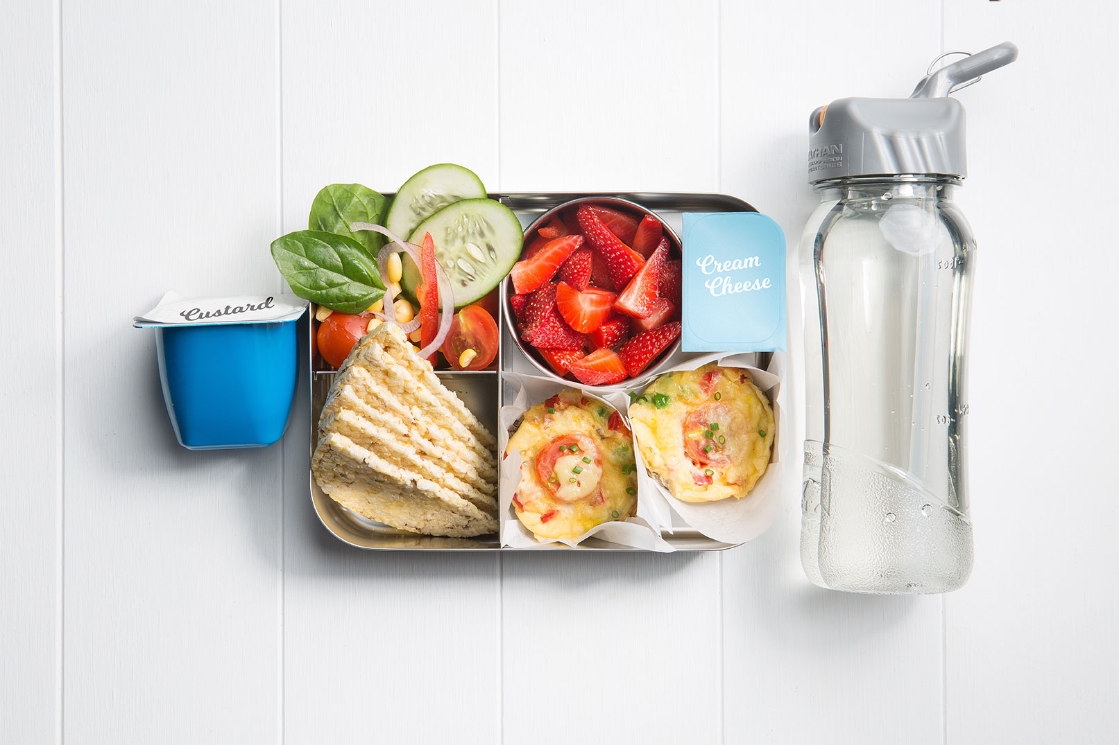 Image of a packed lunch box with mini crustless chicken quiches, mixed salad, corn thins with cream cheese, strawberries, custard tub and a bottle of water on the side