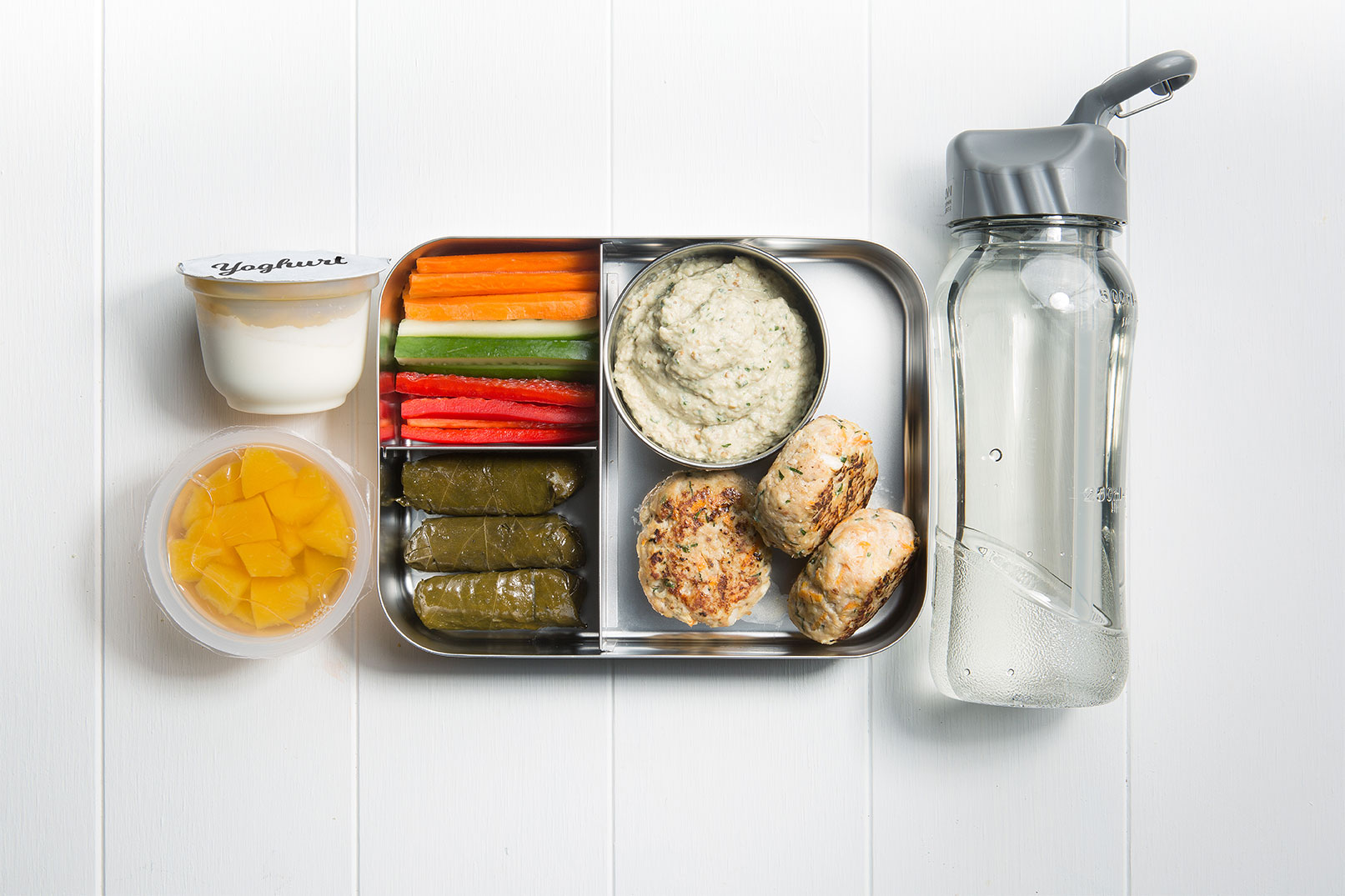 Image of a packed lunch box with dolmades, pork and apple rissoles, babaganoush with vegie sticks and canned fruit, yoghurt and a bottle of water on the side