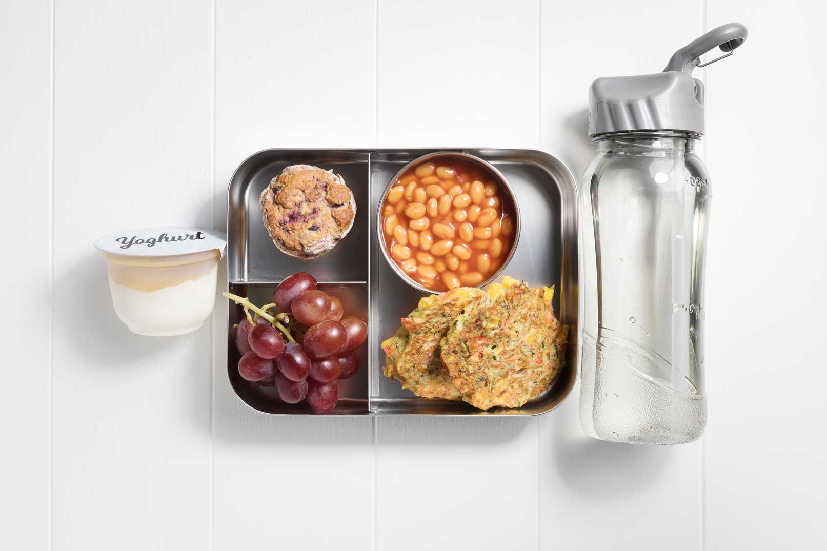 Image of a packed lunch box with two zucchini and corn fritters, baked beans, mixed berry scone, grapes and a yoghurt tub and water bottle on the side