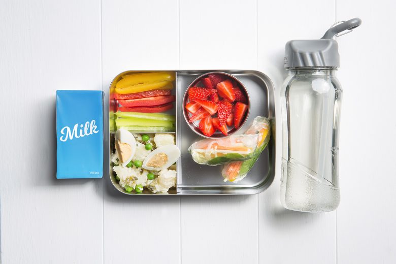 Image of a packed lunch box with potato salad, rice paper rolls, sliced strawberries, three colour capsicum sticks and a plain milk popper and water bottle on the side