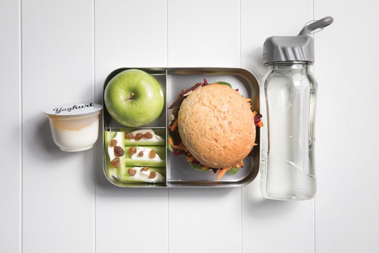 Image of a packed lunch box with a lentil burger, green apple, celery boats and a yoghurt and bottle of water on the side