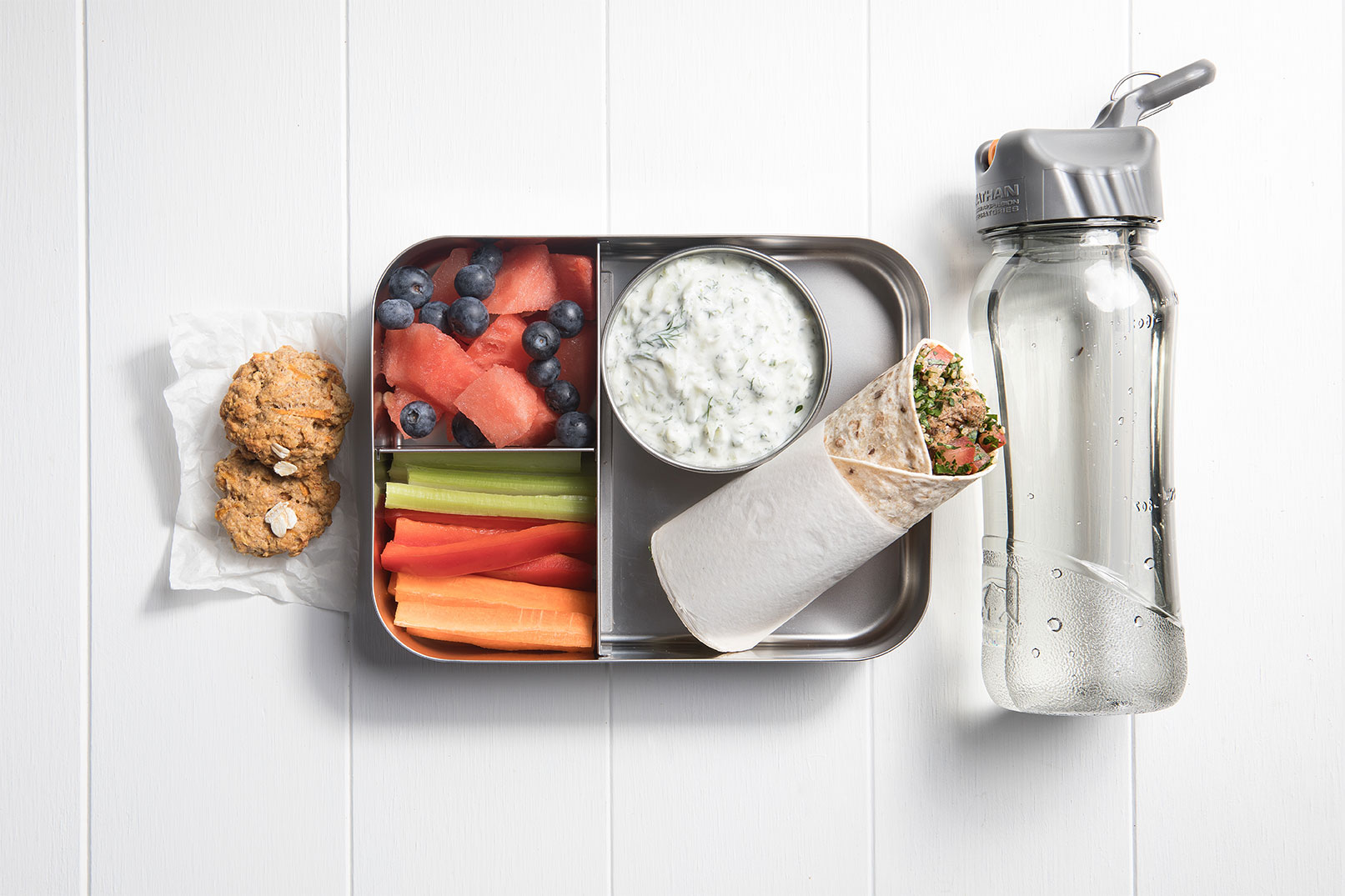 Image of a packed lunch box with beef kofta and tabouli wrap, vegie sticks with tzatziki, watermelon and blueberries, two carrot oast biscuits and a bottle of water on the side