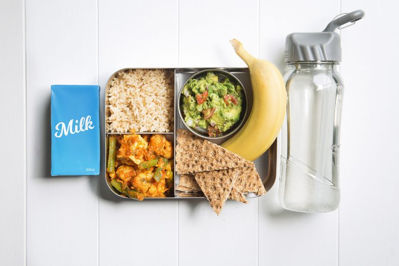 Image of a packed lunch box with tofu korma, brown rice, crackers with guacamole, a banana and a plain milk popper and bottle of water on the side