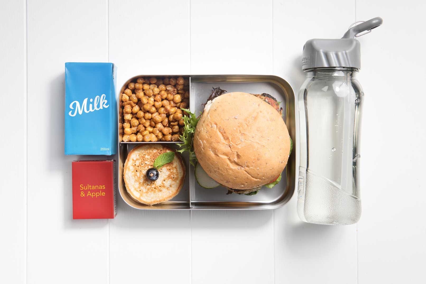 Top view of a packed lunch box with a roll, pikelets, roasted chickpeas, milk popper, container of sultanas and apple and a water bottle.