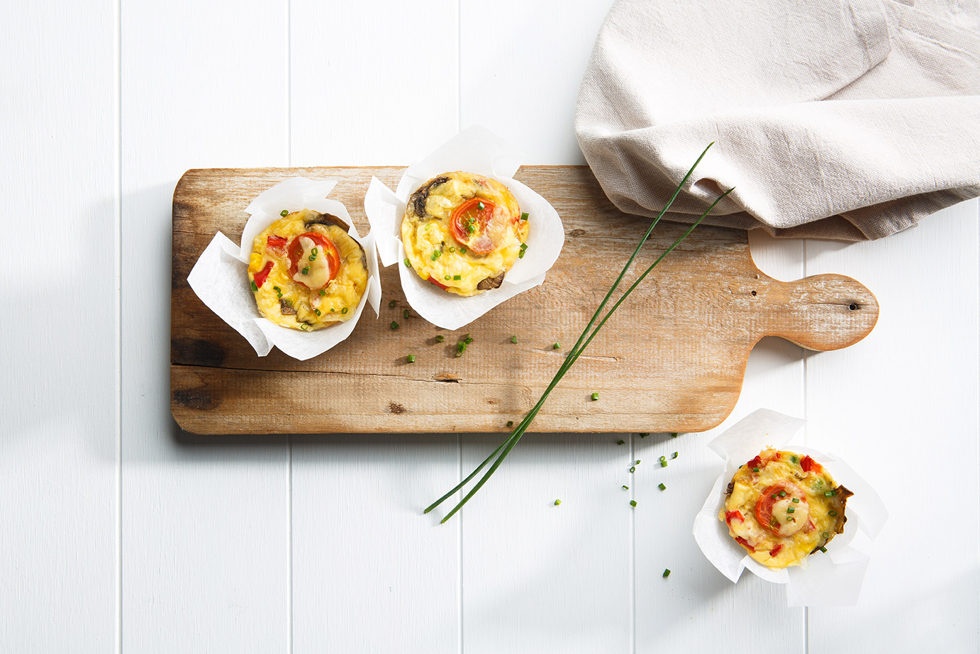 Image of three mini crustless chicken quiches on a wooden chopping board with chives and a white cloth napkin on the side