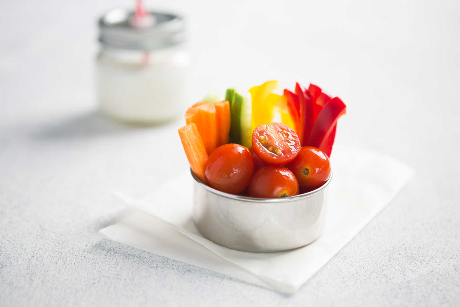 Sliced carrot, cucumber, red capsicum with cherry tomatoes in a silver bowl on a napkin and a container of milk.