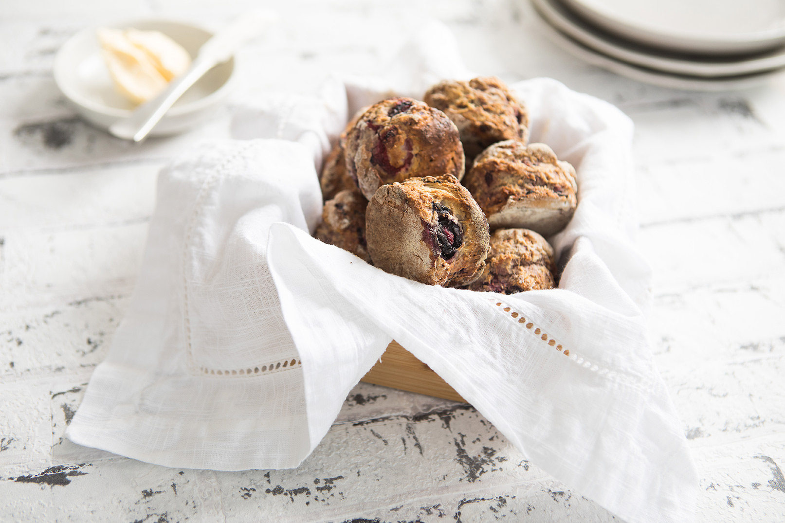 Image of a baked mixed berry scones served on a white cloth napkin sitting in a wooden serving container with a dish of margarine and knife for serving in the background