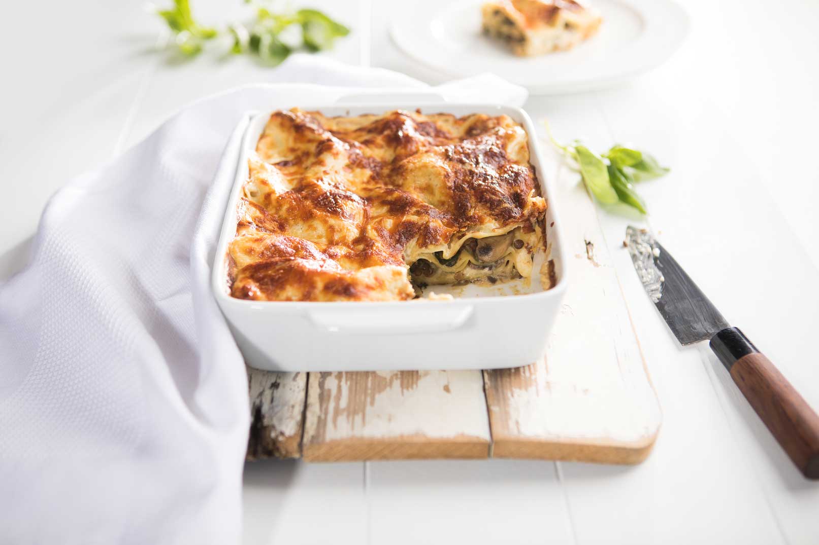 Image of mushroom, spinach and lentil lasagne in a white ceramic dish on a wooden chopping board with basil on the side, a white cloth napkin and a serving knife
