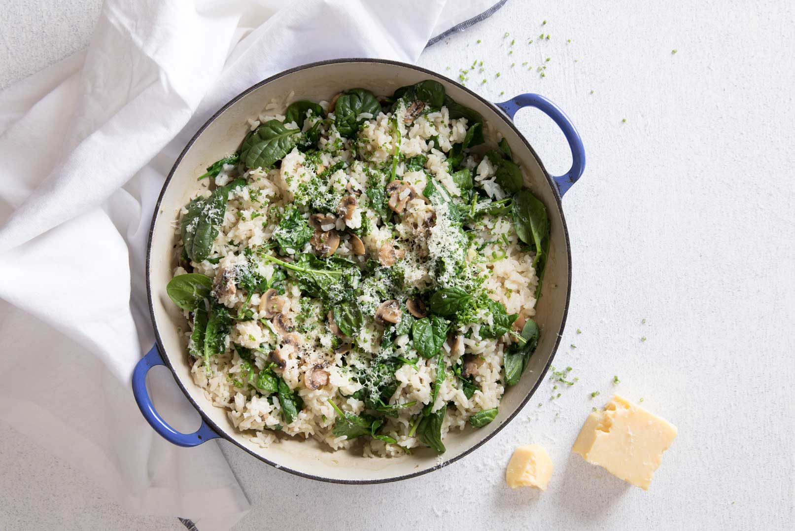 Mushroom risotto bake in a large round oven proof dish with blue handles shot from above and a chunk of parmesan cheese and white cloth napkin on the side.