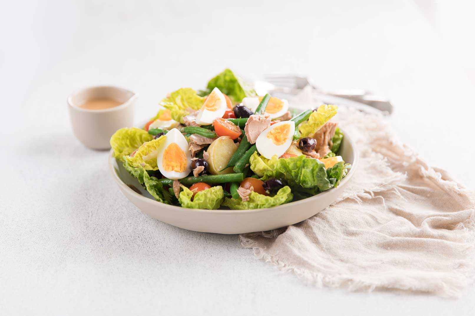 Nicoise salad in a large bowl served on a rustic cloth napkin with dressing on the side and cutlery in the background.