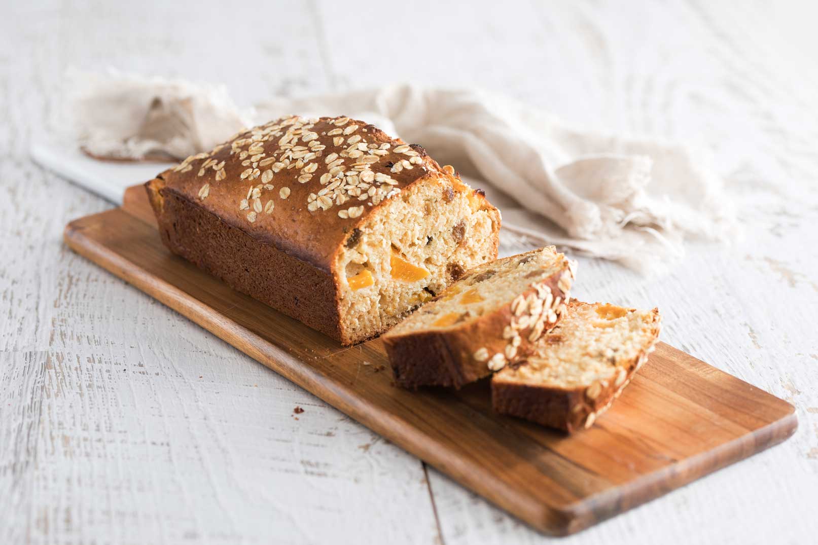 a baked loaf of oat, sultana and peach bread with two slices cut, served on a long wooden serving board with a white cloth napkin in the background