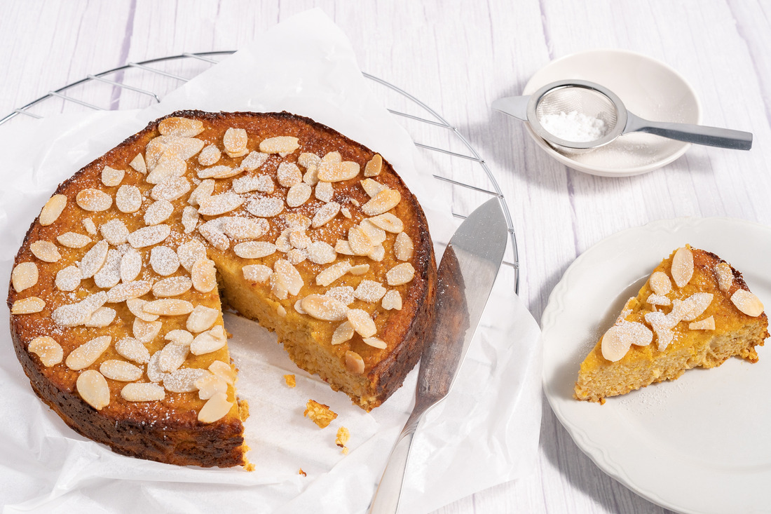 Image of a circular orange and almond cake sitting on a wire rack. The cake has a triangular slice removed and placed on a white side plate beside the cake.