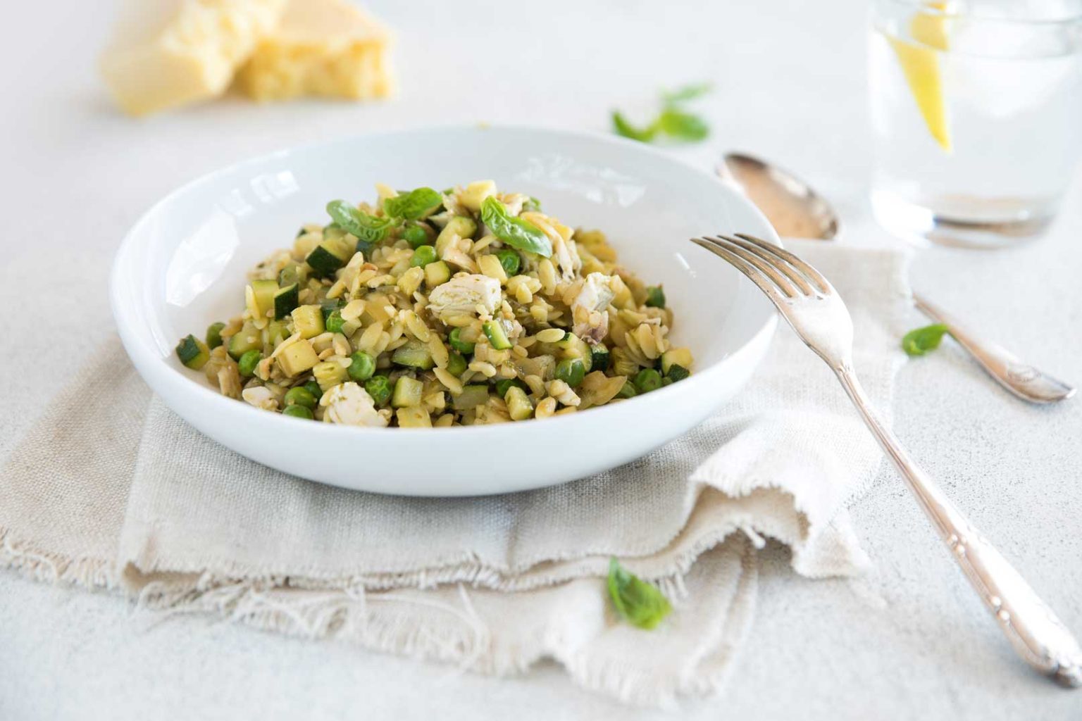 Pea, zucchini and pesto risoni with chicken in a large white bowl with a fork and spoon for serving on the side.