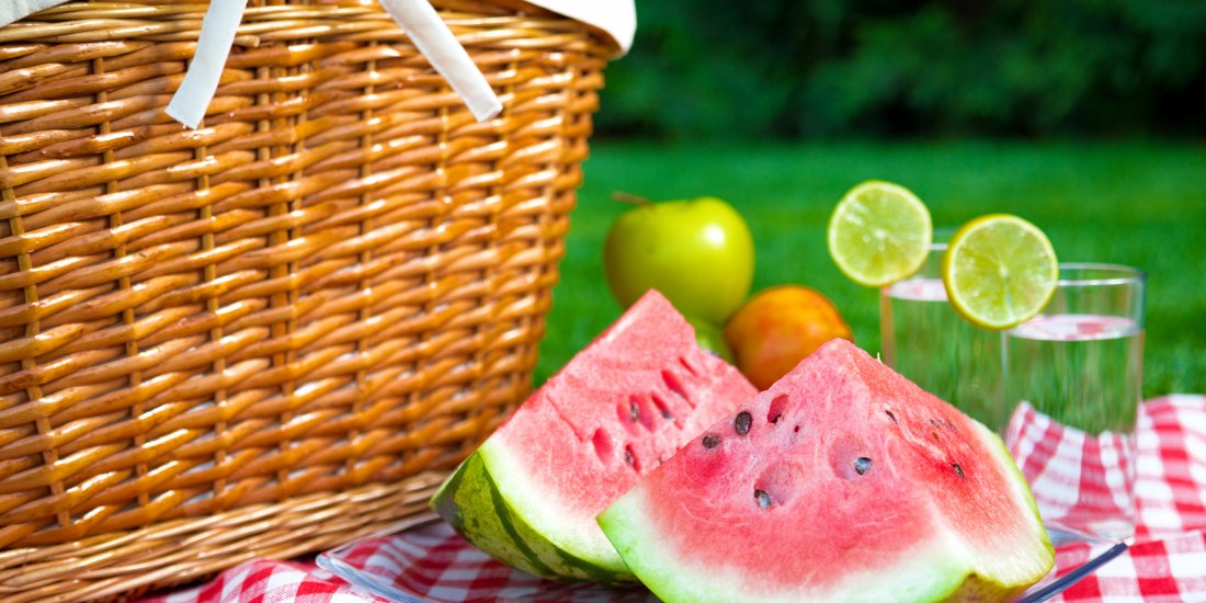 A brown wicker picnic basket with cut up watermelon and water glasses on a red gingham tablecloth