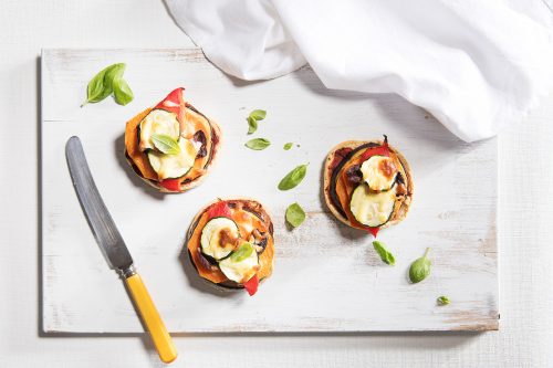 Image of three baked pizza muffins served on a white cutting board with a knife, cloth napkin and scattering of basil on the side
