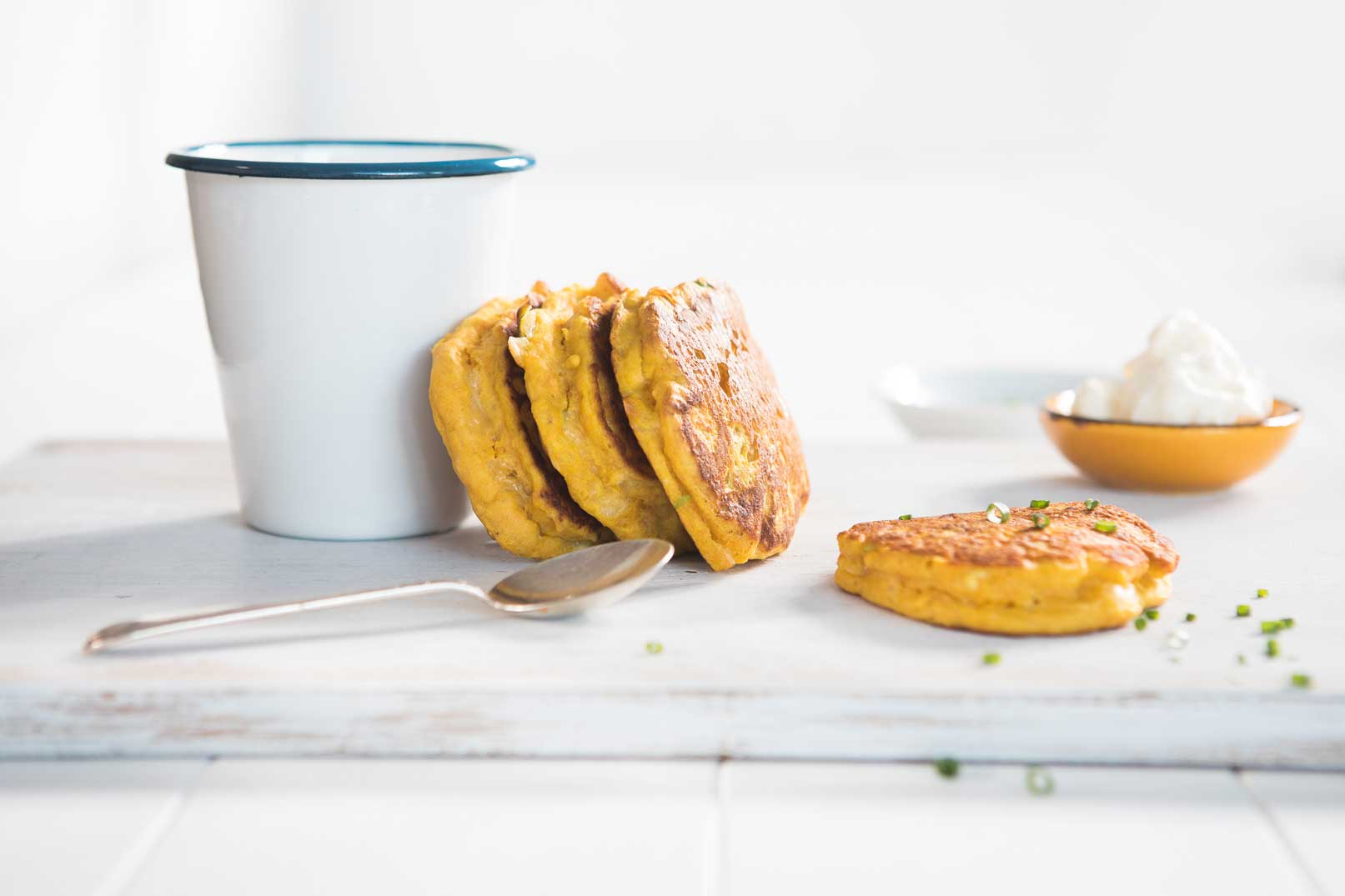 Image of four pumpkin and cheese pikelets with a cup and metal spoon with a side of creme fraiche