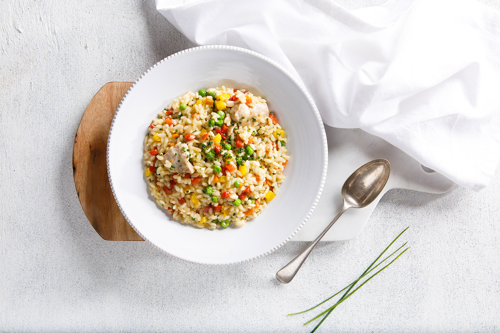 Image of rainbow risotto served in a large white bowl on a wooden cutting board with a napkin, chives and a spoon on the side for serving