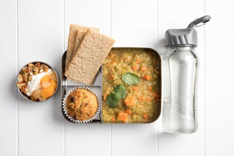 : a compartment metal lunch box packed with red lentil soup, three crispbreads, a carrot and zucchini muffin with a fruit salad parfait to the left and filled water bottle to the right.