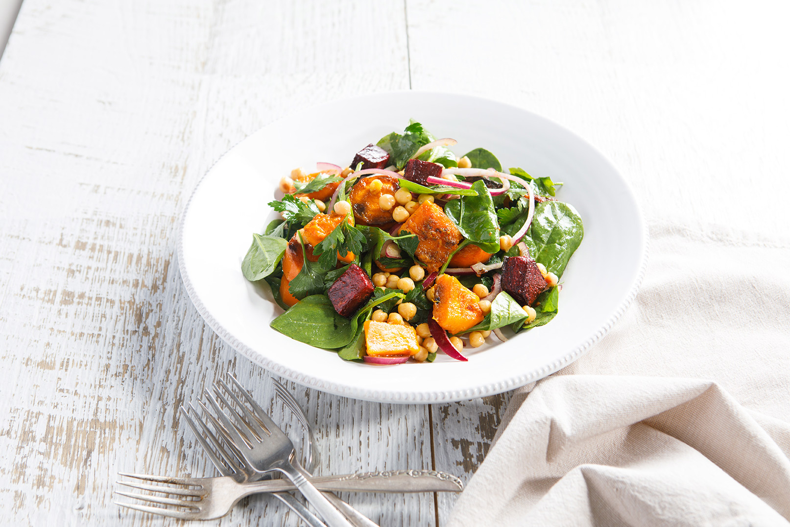 Image of roast vegetable and chickpea salad in a white bowl shot from the side on a wooden table with accompanying forks.