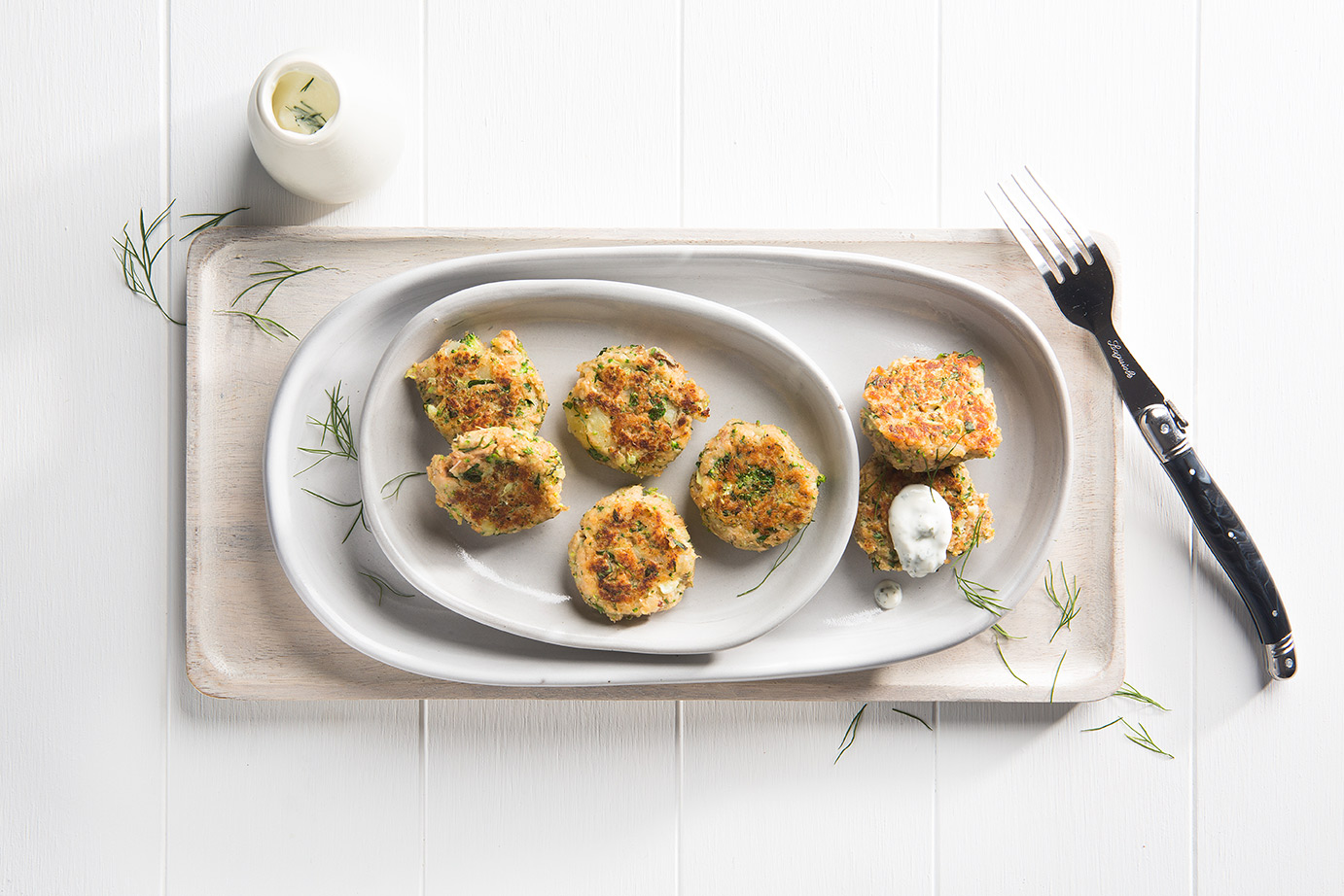 Image of six salmon and potato cakes shot from above on a large stone serving dish on a wooden cutting board with a spoonful of dipping sauce on the side