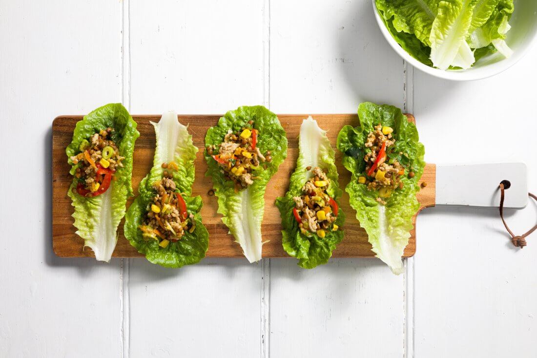 Five lettuce leaves containing meat mixture served on a wooden platter with a bowl of lettuce in the background