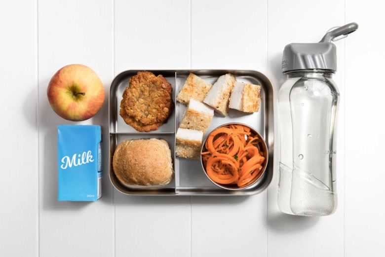 a compartment metal lunch box packed five sesame crusted snapper pieces, small serving dish of carrot salad, wholegrain dinner roll, two Anzac biscuits and an apple and plain milk popper to the left and filled water bottle to the right.
