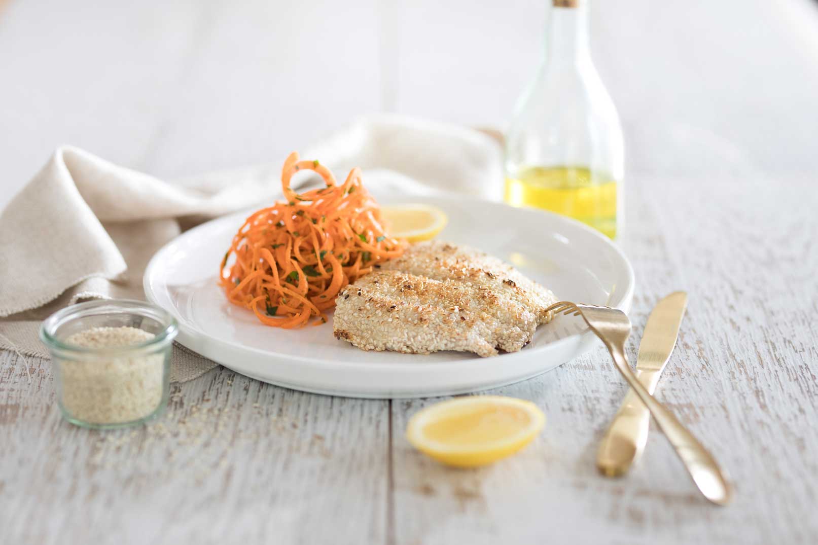 One sesame crusted snapper fillet with carrot salad and a lemon wedge on a white plate served with a gold knife and fork, small glass container of sesame seeds and bottle of dressing and cloth napkin on the side.