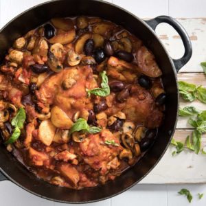 Slow cooker chicken cacciatore recipe in a large black pot with handles served on a white cutting board with a sprinkle of basil on top and the side for serving.