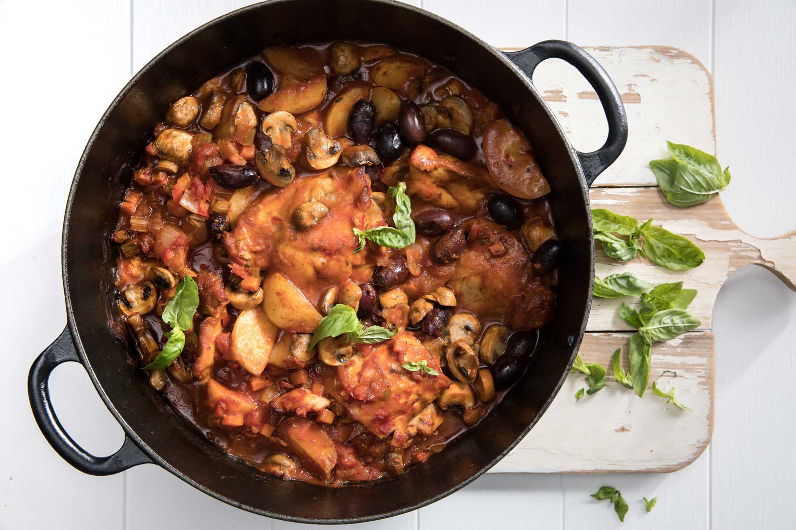Slow cooker chicken cacciatore recipe in a large black pot with handles served on a white cutting board with a sprinkle of basil on top and the side for serving.