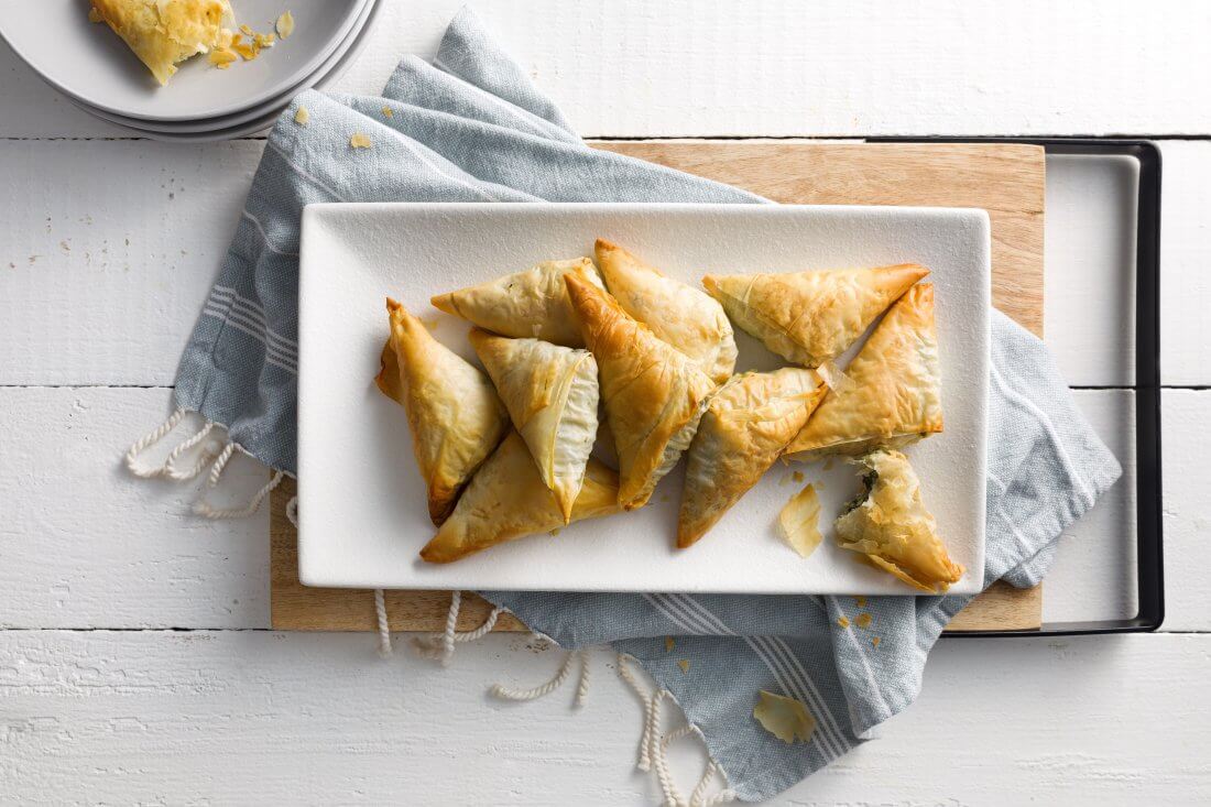 10 small phyllo pastry triangles served on a rectangular white platter on wooden platter with a blue napkin underneath