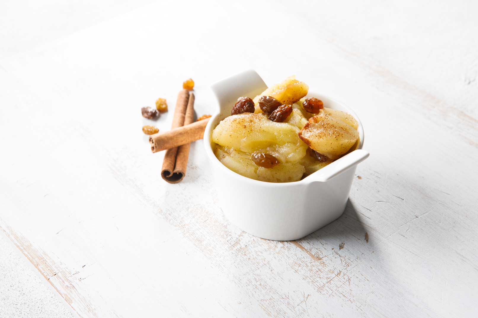 Image of stewed apples topped with sultanas in a white ceramic bowl with two cinnamon sticks on the side.