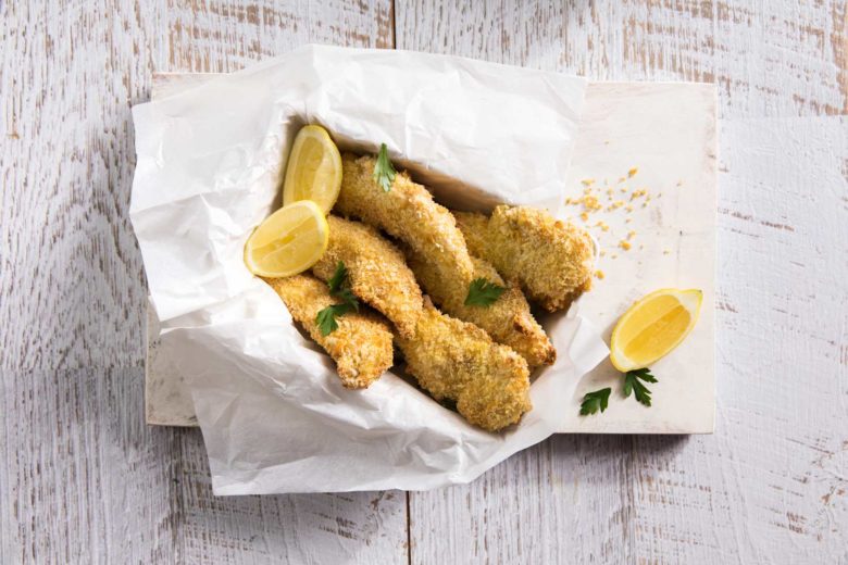 Super crispy chicken fingers arranged on baking paper in a rectangle dish topped with two lemon wedges and parsley leaves served on a white cutting board with a lemon wedge on the side.