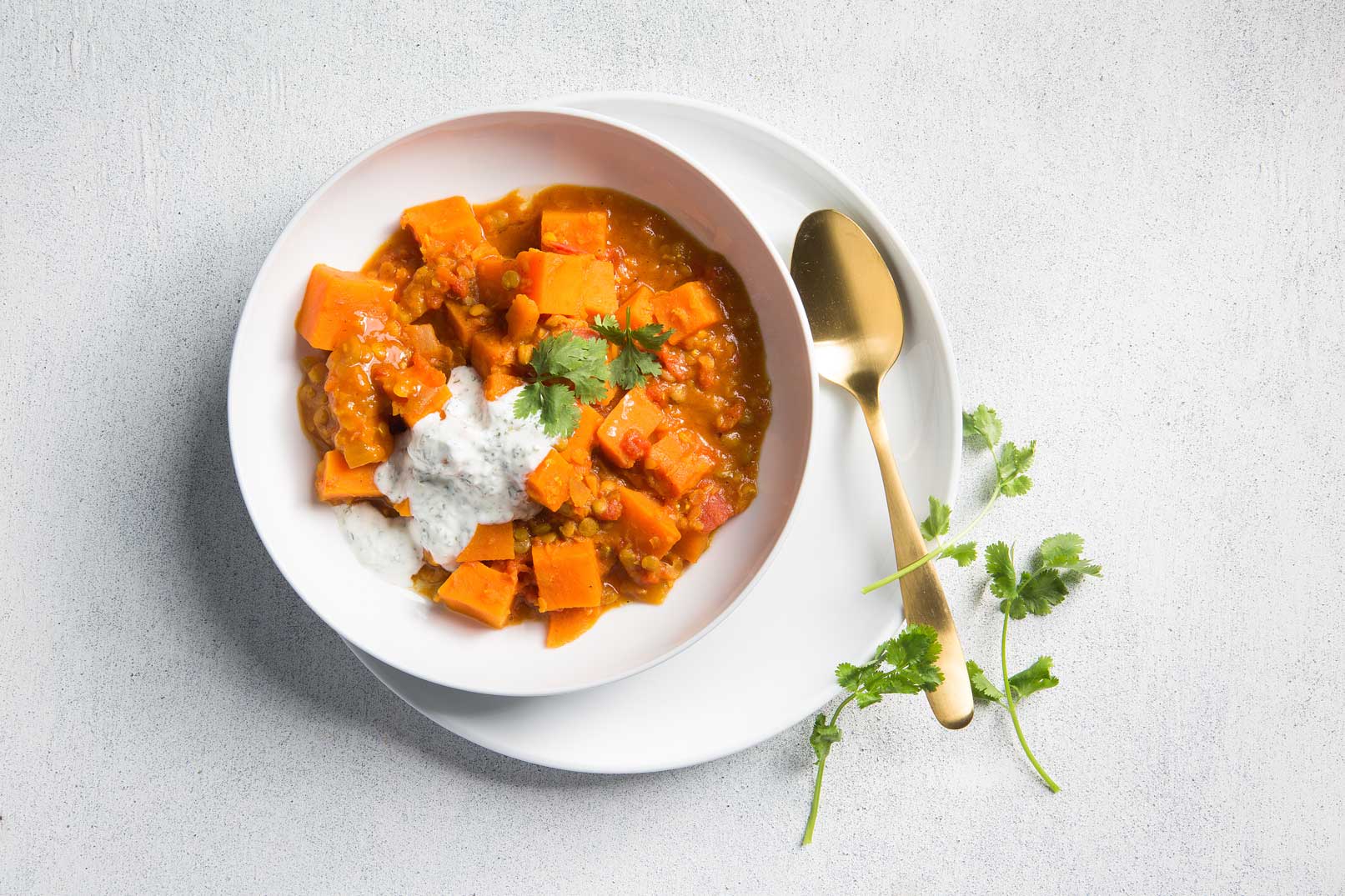 Image of Sweet potato and lentil curry in a white bowl on a white plate topped with yoghurt sauce and coriander and a gold spoon for serving