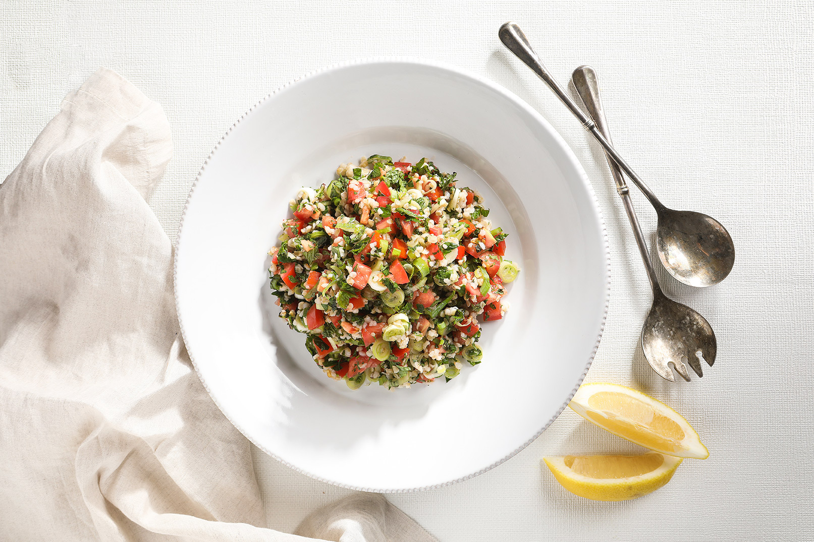 Image of tabouli on a white plate shot from above with lemon wedges and serving spoons.