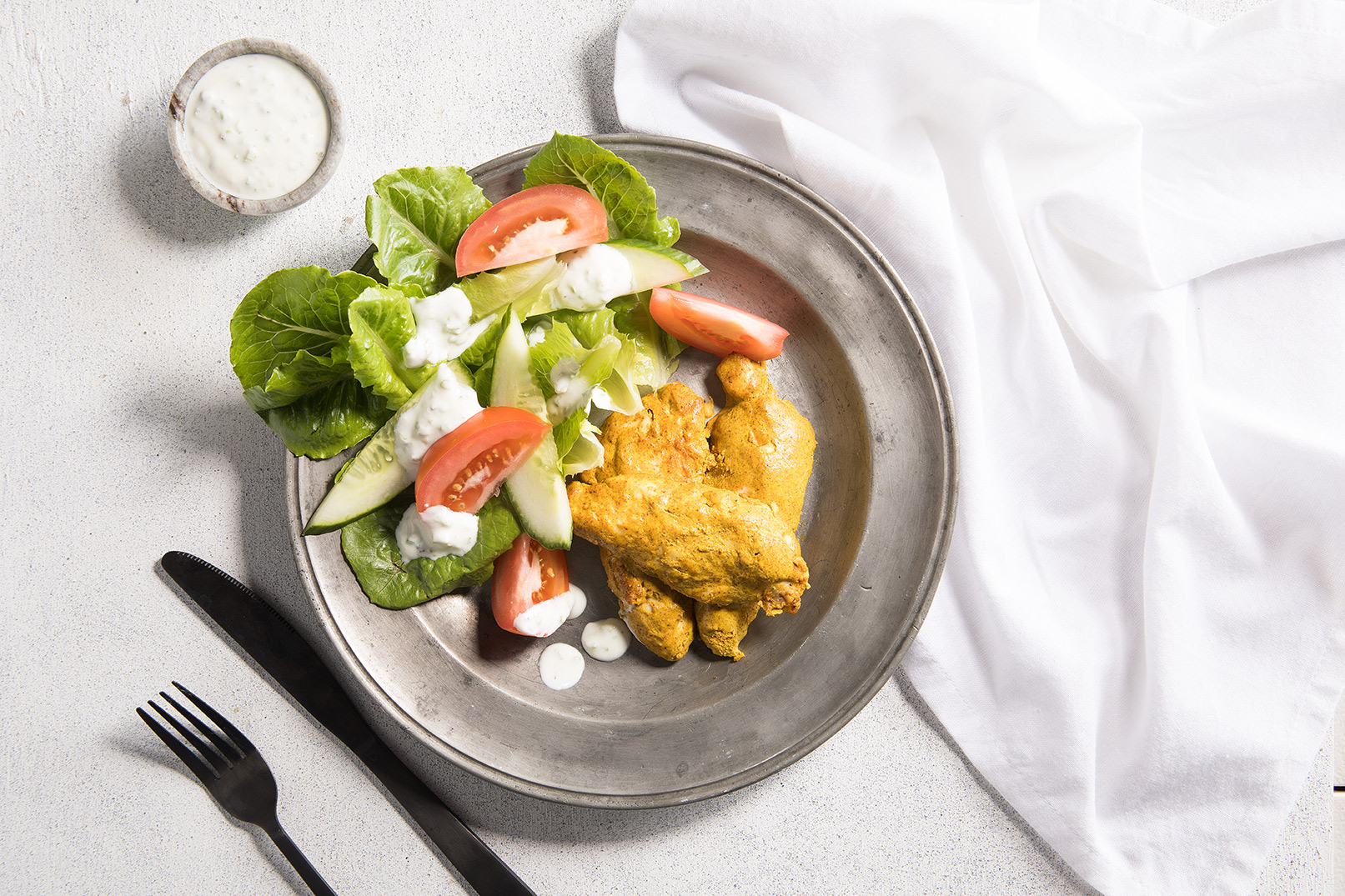 Image of tandoori chicken with salad on a silver bowl with a side of dressing, silver knife and fork and white cloth napkin.