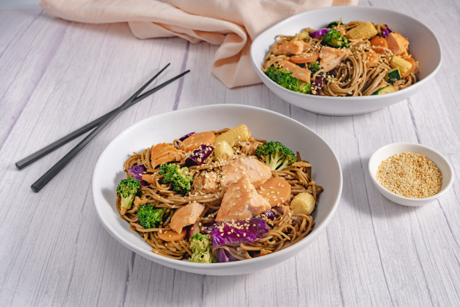 Two white bowls on a white table. Within the bowls there is a colourful teriyaki salmon and noodle stirfry.