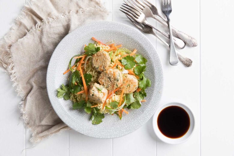 Thai chicken meatballs on a bed of fresh Asian salad served on a large white plate with dipping sauce, cutlery and cloth napkin on the side.