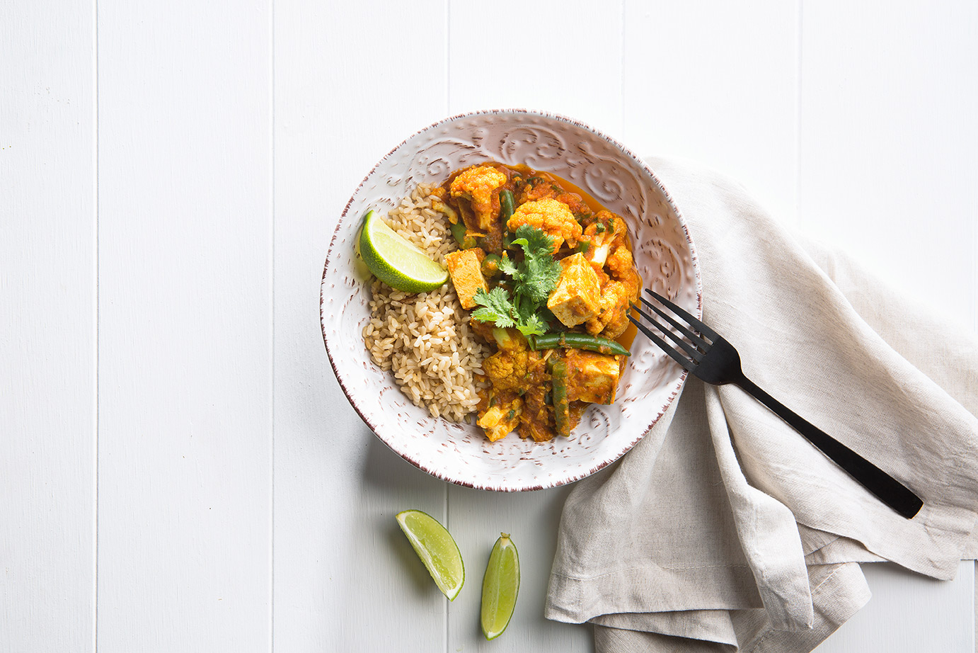 Image of cooked tofu korma recipe in a white bowl with fork, cloth napkin and lime wedges on the side for serving 