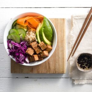 round white bowl containing tofu, brown rice, avocado, cucumber, carrot and red cabbage on a wooden platter with wooden chopsticks and a small bowl of soy sauce on the right