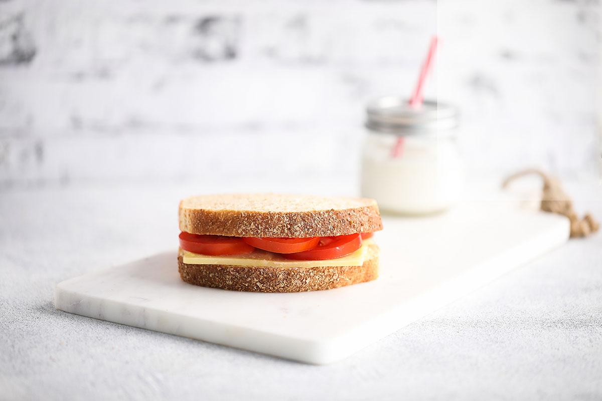 Image of a tomato-cheese-sandwich served on a white cutting board with a glass jar of milk with straw on the side