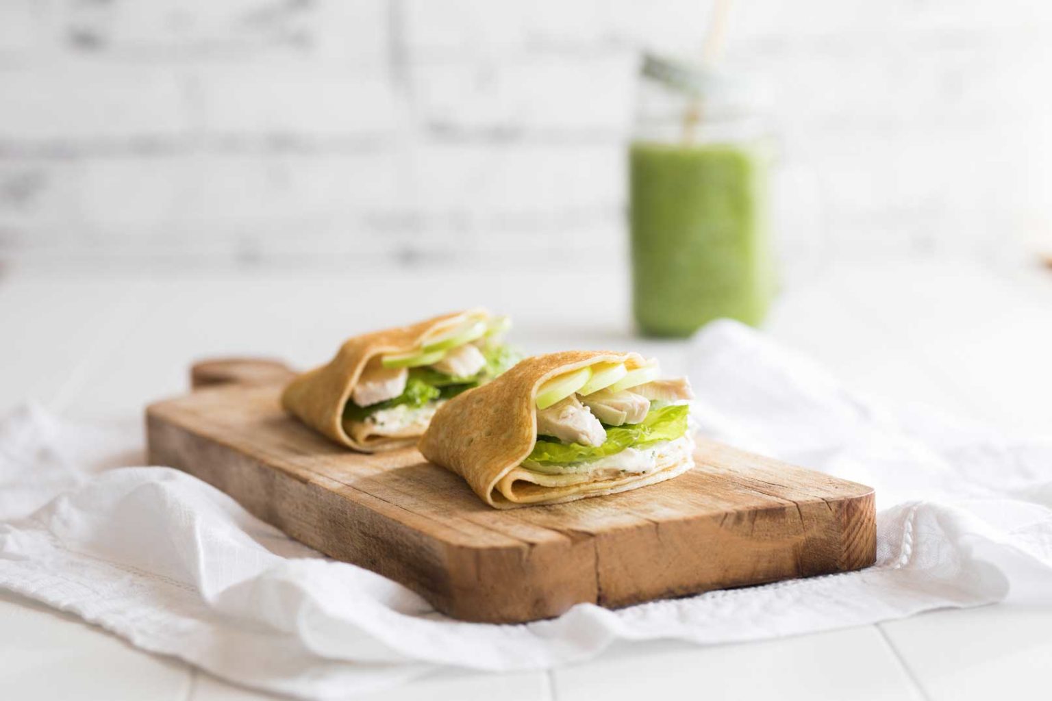 Two turkey, apple and avocado crepes on a wooden serving board sitting on white cloth napkin with a green smoothie in a glass jar in the background.