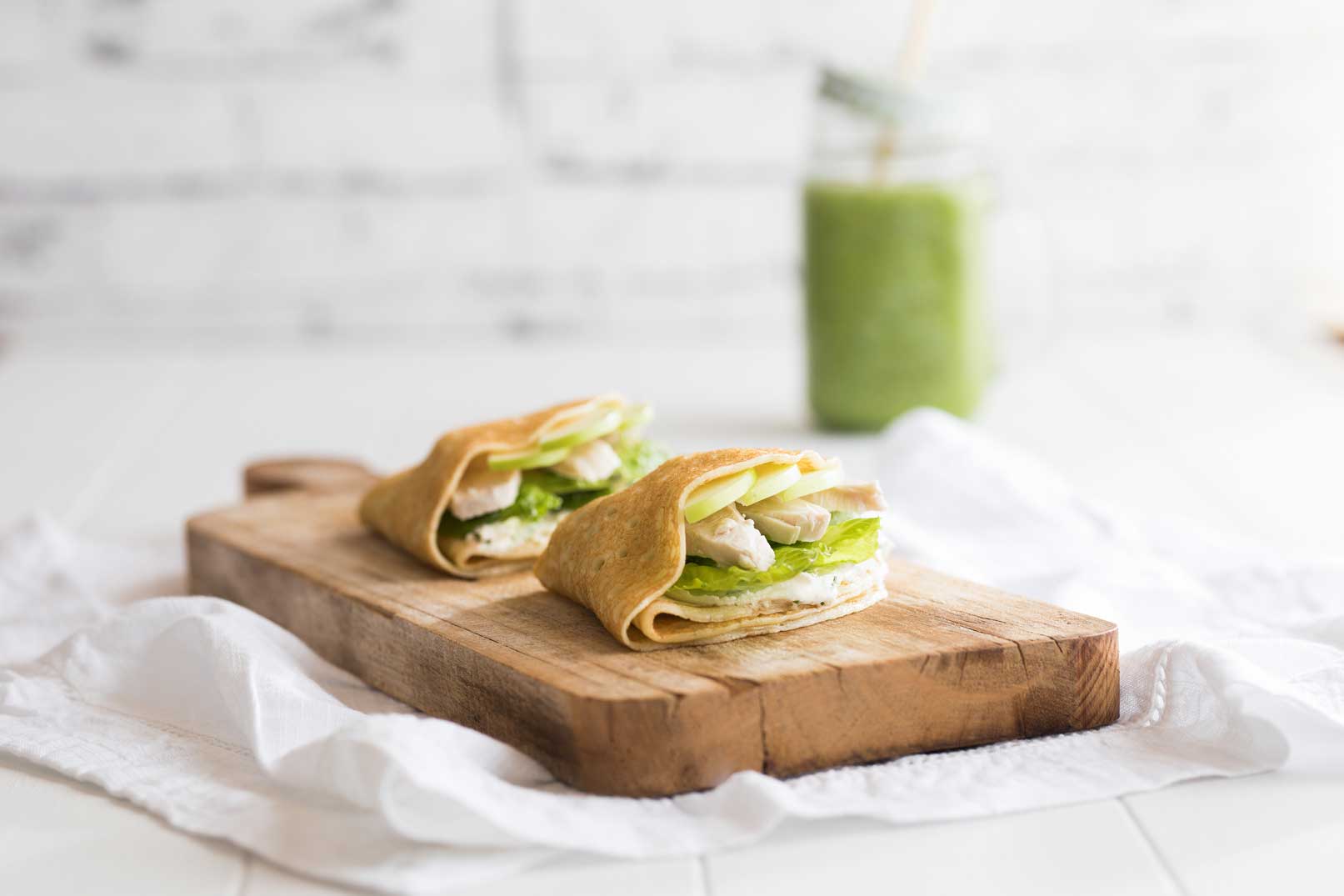 Two turkey, apple and avocado crepes on a wooden serving board sitting on white cloth napkin with a green smoothie in a glass jar in the background.