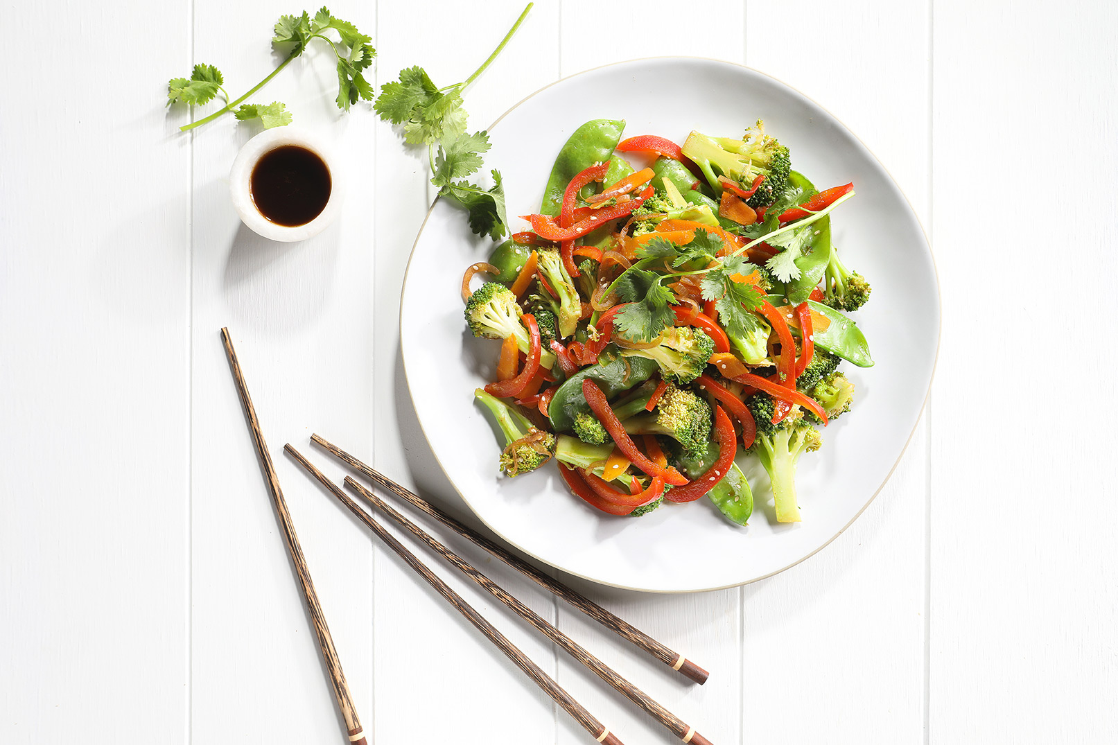 Image of stir fry vegetables on a large white plate with herbs, dressing and wooden chopsticks on the side