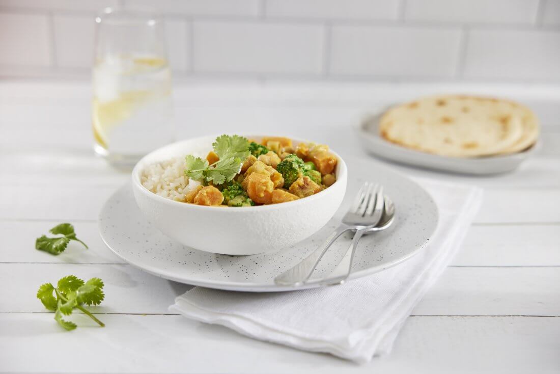 A white bowl containing vegetable curry with a spoon and fork and a glass of water and naan bread in the background